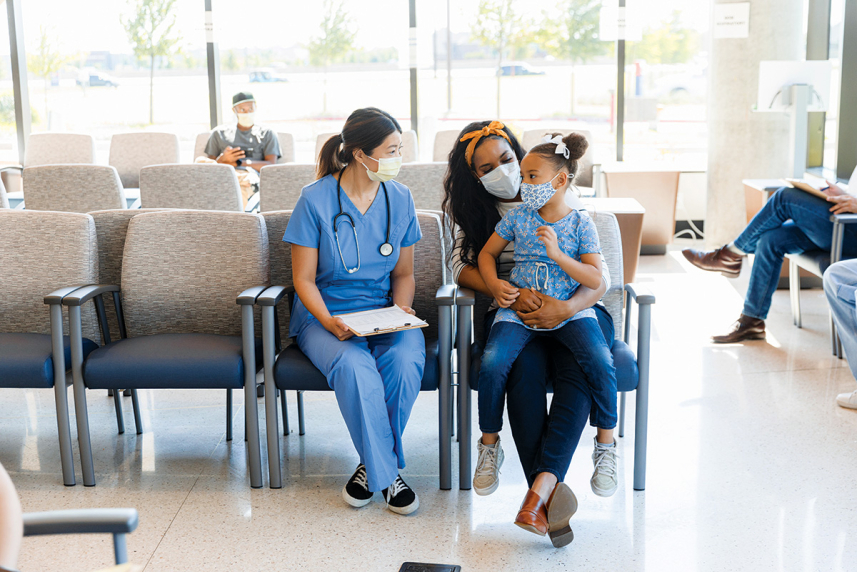 A child sits on an adult's lap next to a doctor. They are all wearing masks.