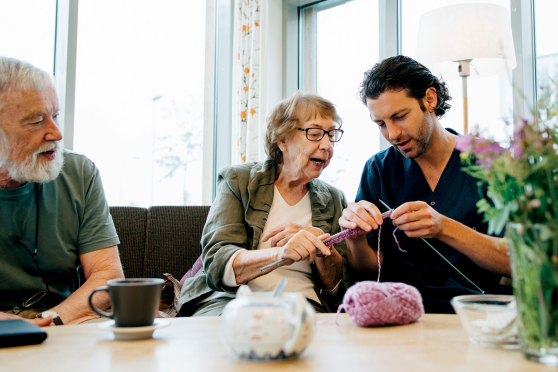 An older man looking at an older woman teaching a nurse to knit.